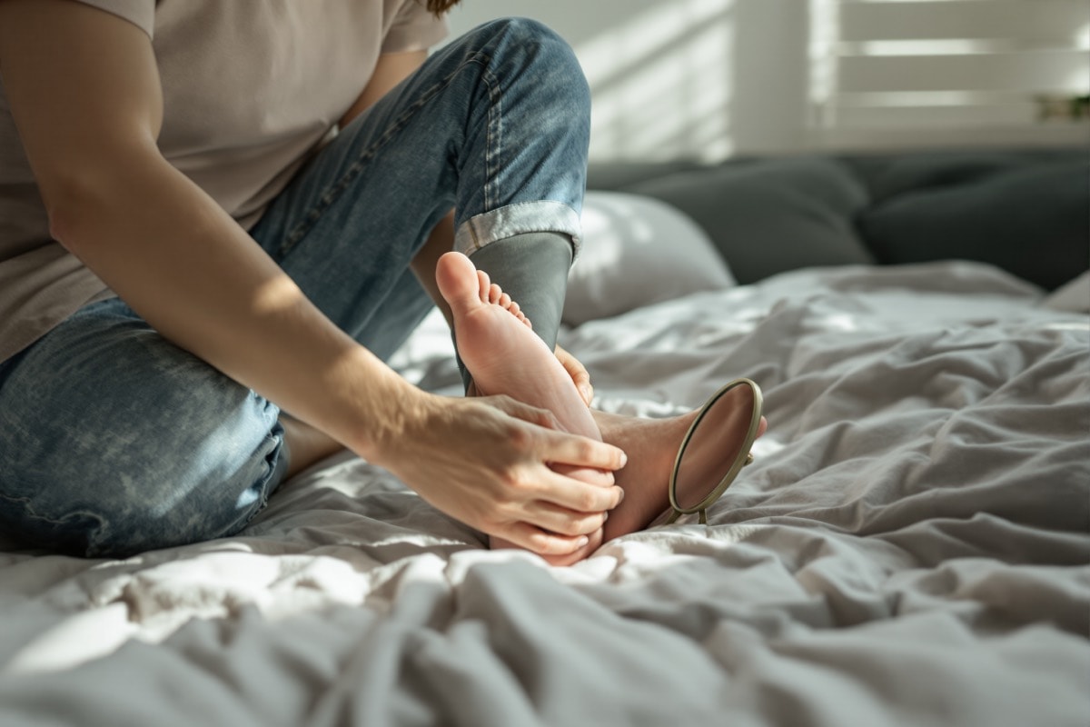 Person seated on a bed inspecting the sole of their own foot with a hand mirror